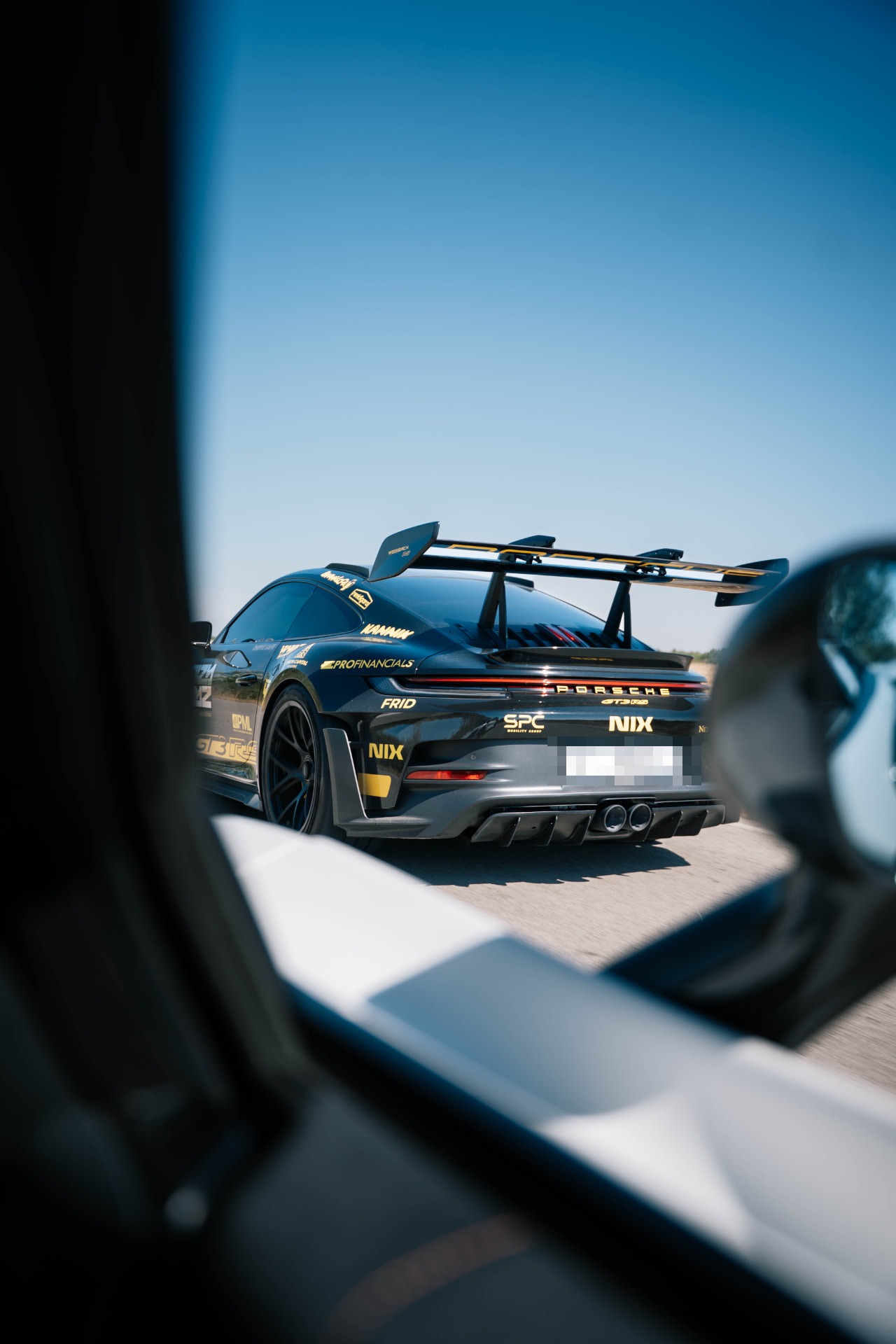 Black Porsche 911 GT3 RS carrying gold sponsor graphics, filmed from a chase car