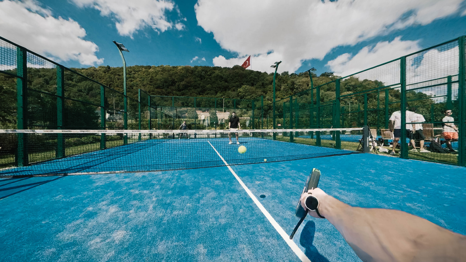 A padel match on an outdoor court, green hills and a Turkish flag in the distance
